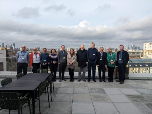 Image shows a group of adult men and women, standing on a roof terrace against a blue and slightly cloudy sky.